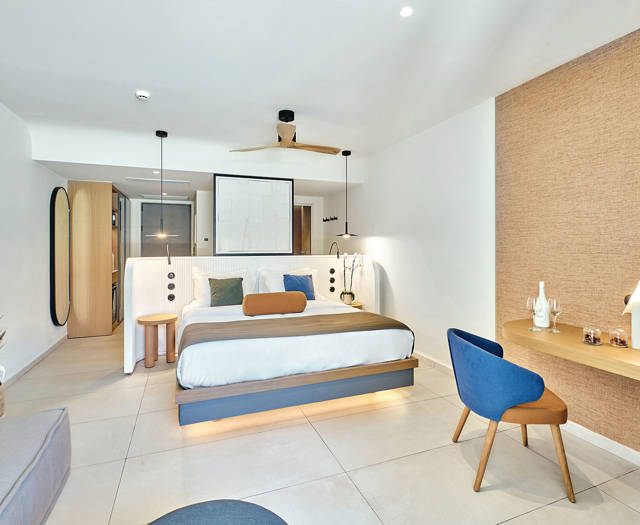 Bright junior suite with double bed, sofa, blue and green chairs, and a desk