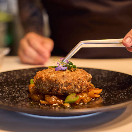 Chef carefully placing edible flowers on a crusted meat dish served with sautéed vegetables on a black speckled plate.