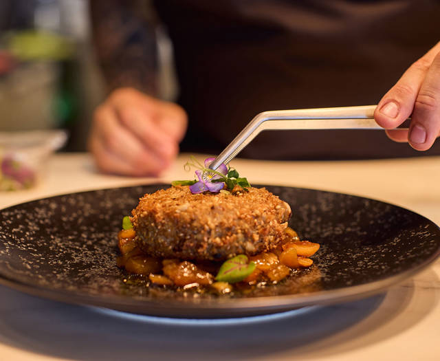Chef carefully placing edible flowers on a crusted meat dish served with sautéed vegetables on a black speckled plate.
