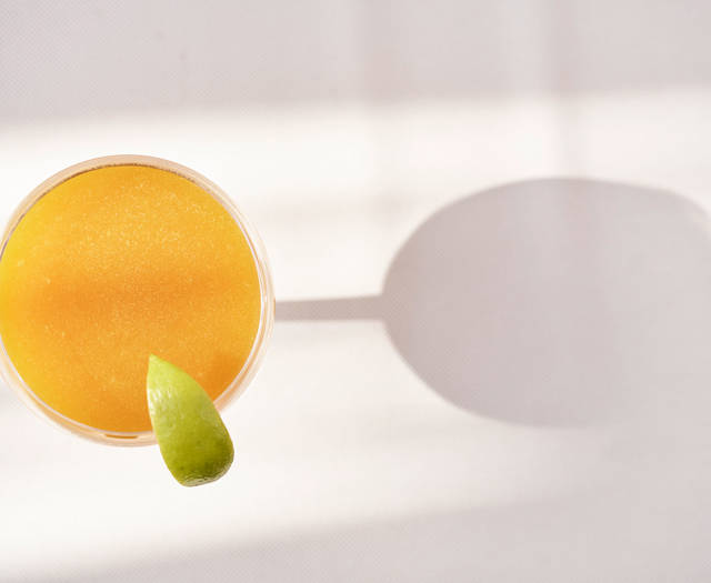 Top-down view of a bright yellow cocktail garnished with a lime wedge, casting a shadow on a white surface.