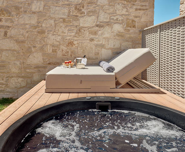 Outdoor hydromassage tub facing a sun lounger with drinks and fresh fruit in a private garden setting.