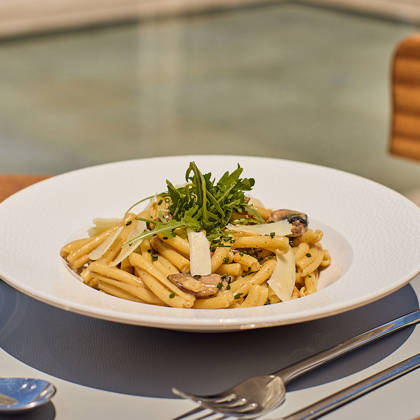 Creamy pasta with mushrooms, shaved cheese, and fresh greens served in an outdoor dining setting.
