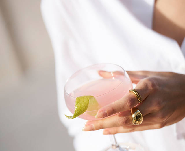 Close-up of a woman in a white shirt holding a pink cocktail garnished with a lime twist