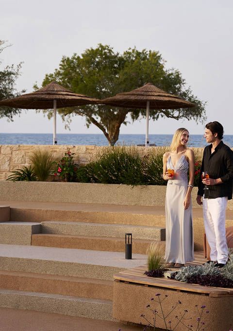 Couple dressed elegantly, standing and enjoying drinks in an outdoor terraced lounge area overlooking the sea with trees and sun umbrellas