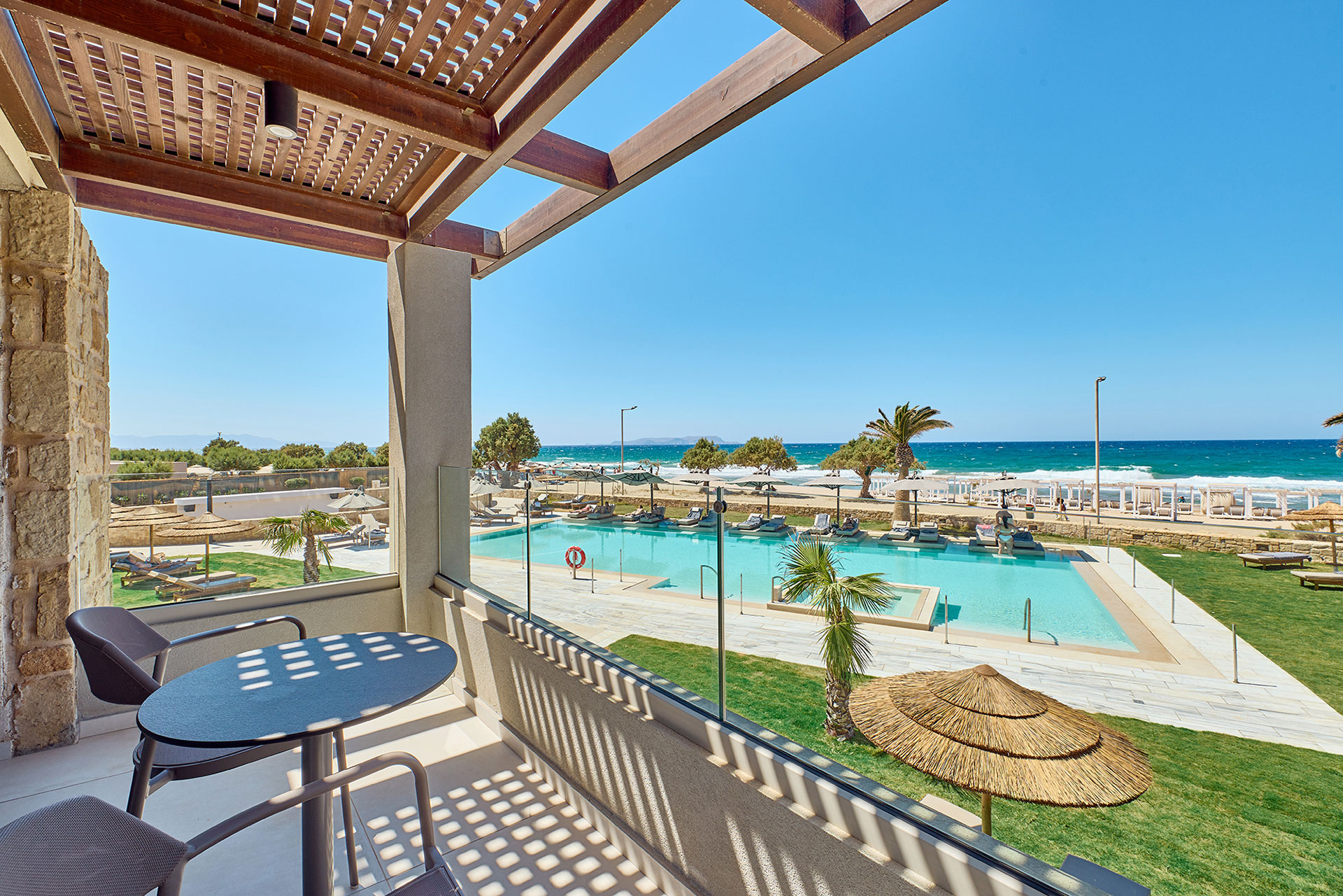 Balcon sous une pergola en bois, avec table et chaises donnant sur la piscine, la plage et la mer.