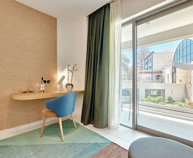 Junior suite desk corner with chair, wine setup, and balcony view of modern architecture.