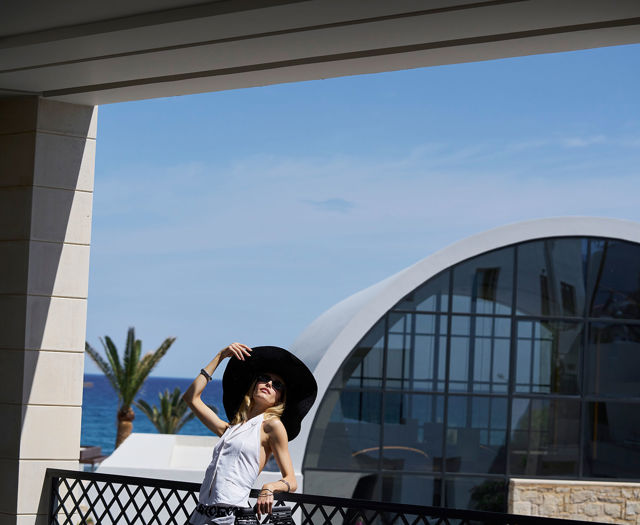 Woman in white outfit and black sunhat posing in front of a curved glass building with sea view