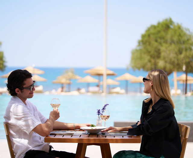Couple dining by the pool with a sea view and parasols in the background.