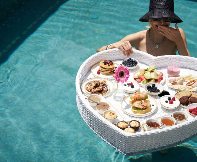 Woman enjoying a floating heart-shaped breakfast tray in a pool filled with various dishes and fruit