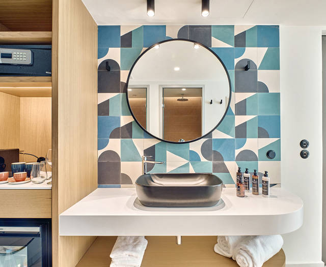 Modern bathroom with bold geometric tiles, round mirror, and open shelving with minibar and safe.