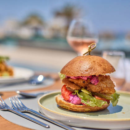 Crispy chicken burger with fresh vegetables and pink sauce, served outdoors with rosé wine by the sea.