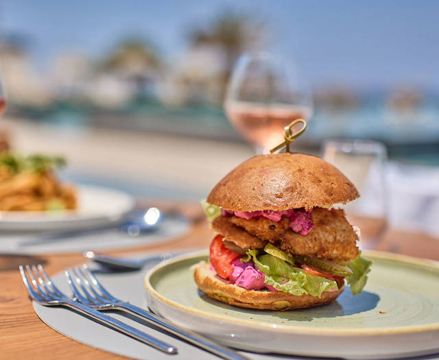 Crispy chicken burger with fresh vegetables and pink sauce, served outdoors with rosé wine by the sea.