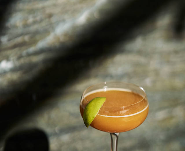 Cocktail glass filled with an amber drink garnished with a lime wedge, set on a wooden table with soft natural lighting.