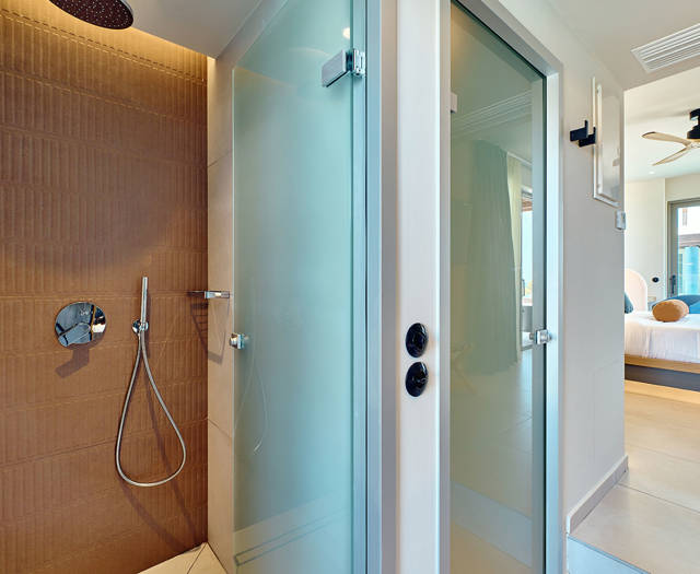 Modern bathroom with brown-tiled walk-in shower and adjacent frosted glass doors, leading to bedroom with sea view.