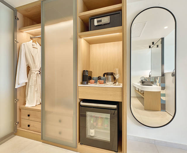 An open wooden wardrobe and wooden shelves with minibar and safe next to a modern mirror.