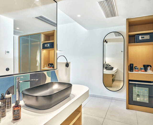 Sleek black sink basin on a white counter with colorful geometric tile backsplash and wooden shelves with minibar and safe