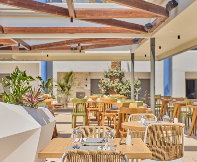 Sunny outdoor restaurant area with wooden tables, modern decor, and a partially open roof structure.