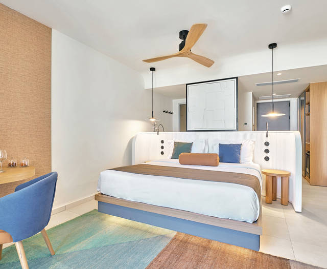 Bright junior suite with double bed, blue chair and a desk, and colorful area rug.