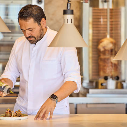 Chef adding sauce to plated Greek wraps in an open kitchen with a vertical rotisserie in the background.