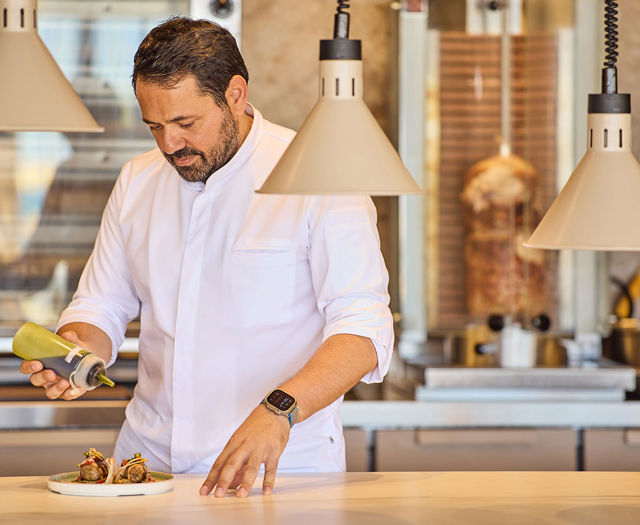 Chef adding sauce to plated Greek wraps in an open kitchen with a vertical rotisserie in the background.