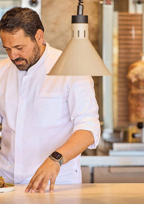 Chef adding sauce to plated Greek wraps in an open kitchen with a vertical rotisserie in the background.