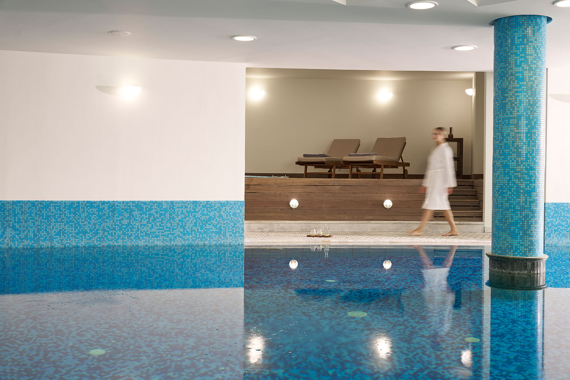 Guest in bathrobe walking by indoor spa pool with mosaic tiles.