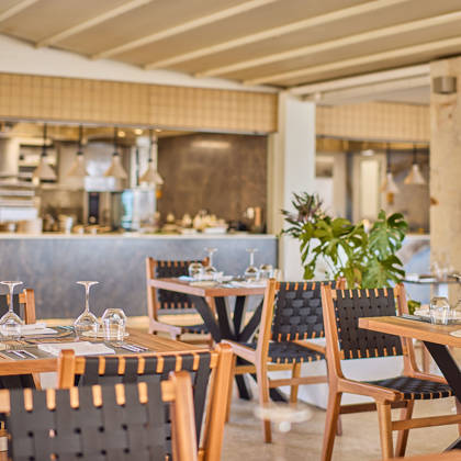 Elegant steakhouse dining area with wooden tables, black woven chairs, and an open kitchen in the background.