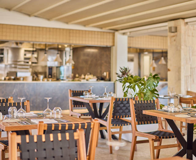 Elegant steakhouse dining area with wooden tables, black woven chairs, and an open kitchen in the background.