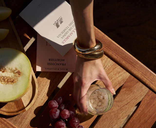 Hand holding iced drink next to melon, grapes, and books on wooden table.
