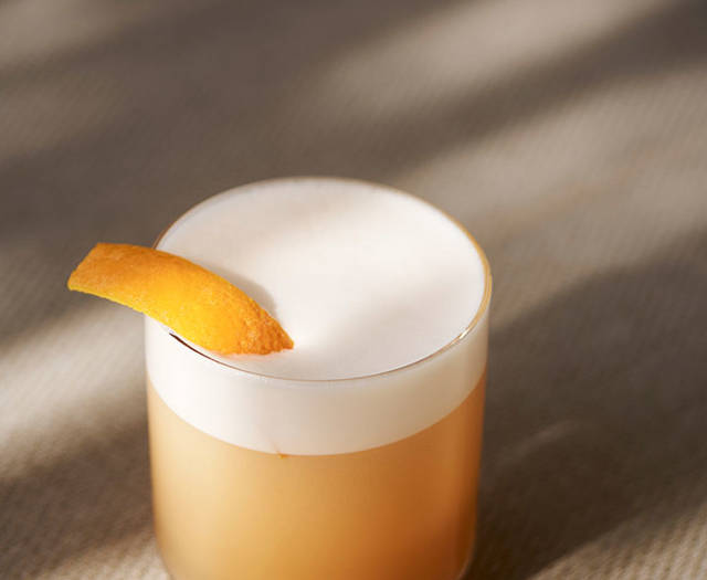 Cocktail with a frothy top and an orange peel garnish, set on a textured surface with soft natural light.