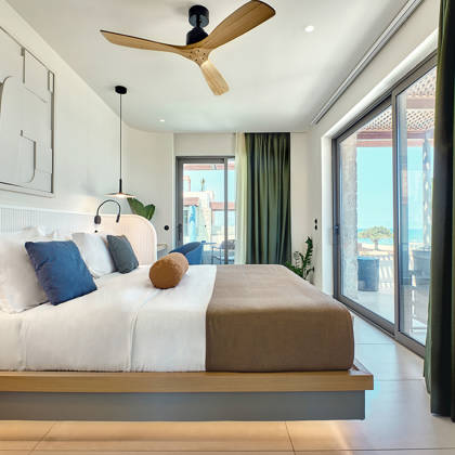Modern bedroom with a large bed, wooden ceiling fan, and sea view through glass doors and windows.