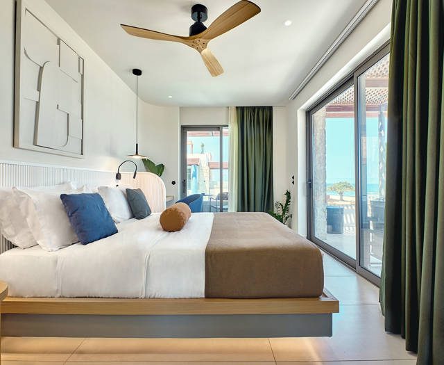 Modern bedroom with a large bed, wooden ceiling fan, and sea view through glass doors and windows.