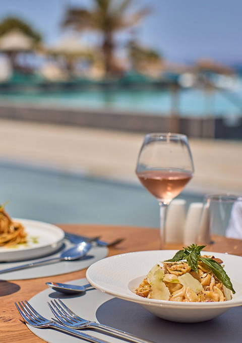 Elegant pasta dishes and rosé wine served at a seaside table with views of a luxurious pool and palm trees.