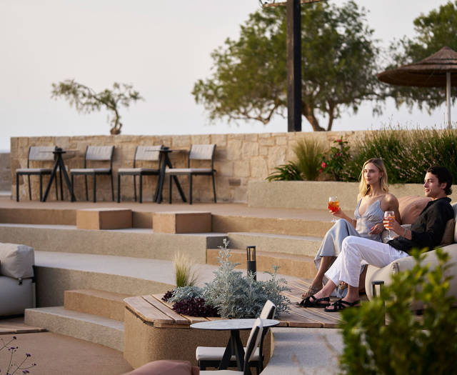 Couple enjoying drinks while seated on an outdoor lounge overlooking terraced steps and greenery near the sea.