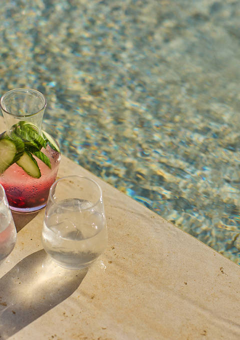 Refreshing pitcher of cucumber and berry-infused water with two glasses, placed poolside in bright sunlight.