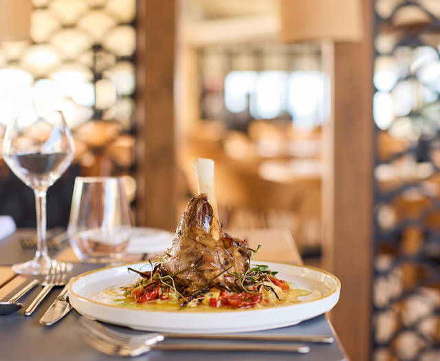Braised lamb shank served on creamy polenta with vegetables, elegantly plated in a sunlit restaurant setting.