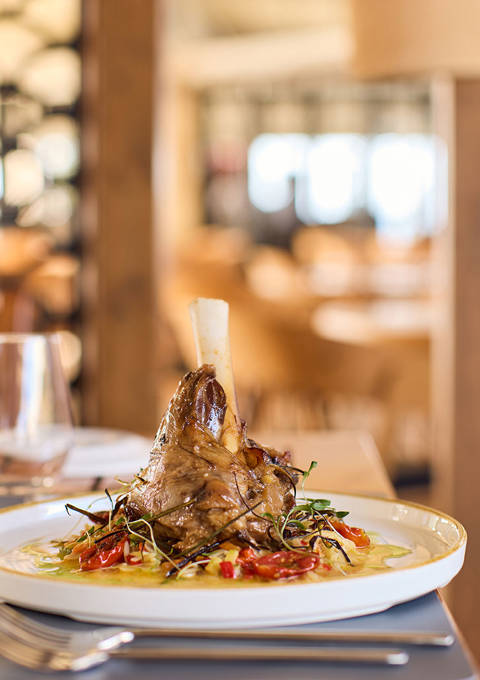 Braised lamb shank served on creamy polenta with vegetables, elegantly plated in a sunlit restaurant setting.