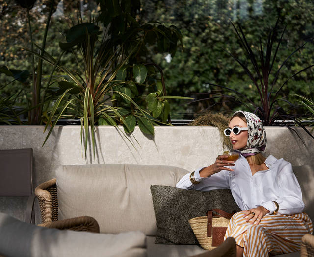 Stylish woman wearing white sunglasses and a headscarf, seated on a cushioned sofa surrounded by plants, sipping a cocktail.