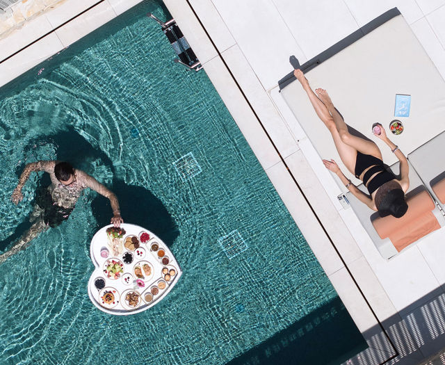 Woman sunbathing by the pool while a man swims with a floating breakfast tray.