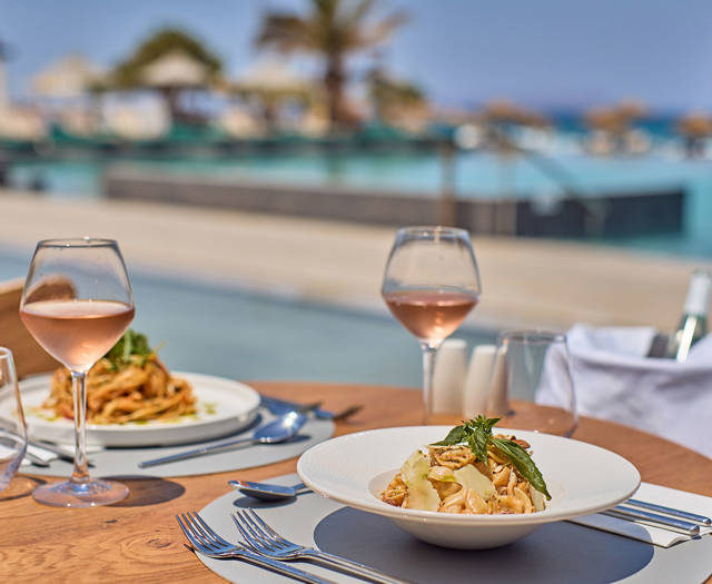 Elegant pasta dishes and rosé wine served at a seaside table with views of a luxurious pool and palm trees.
