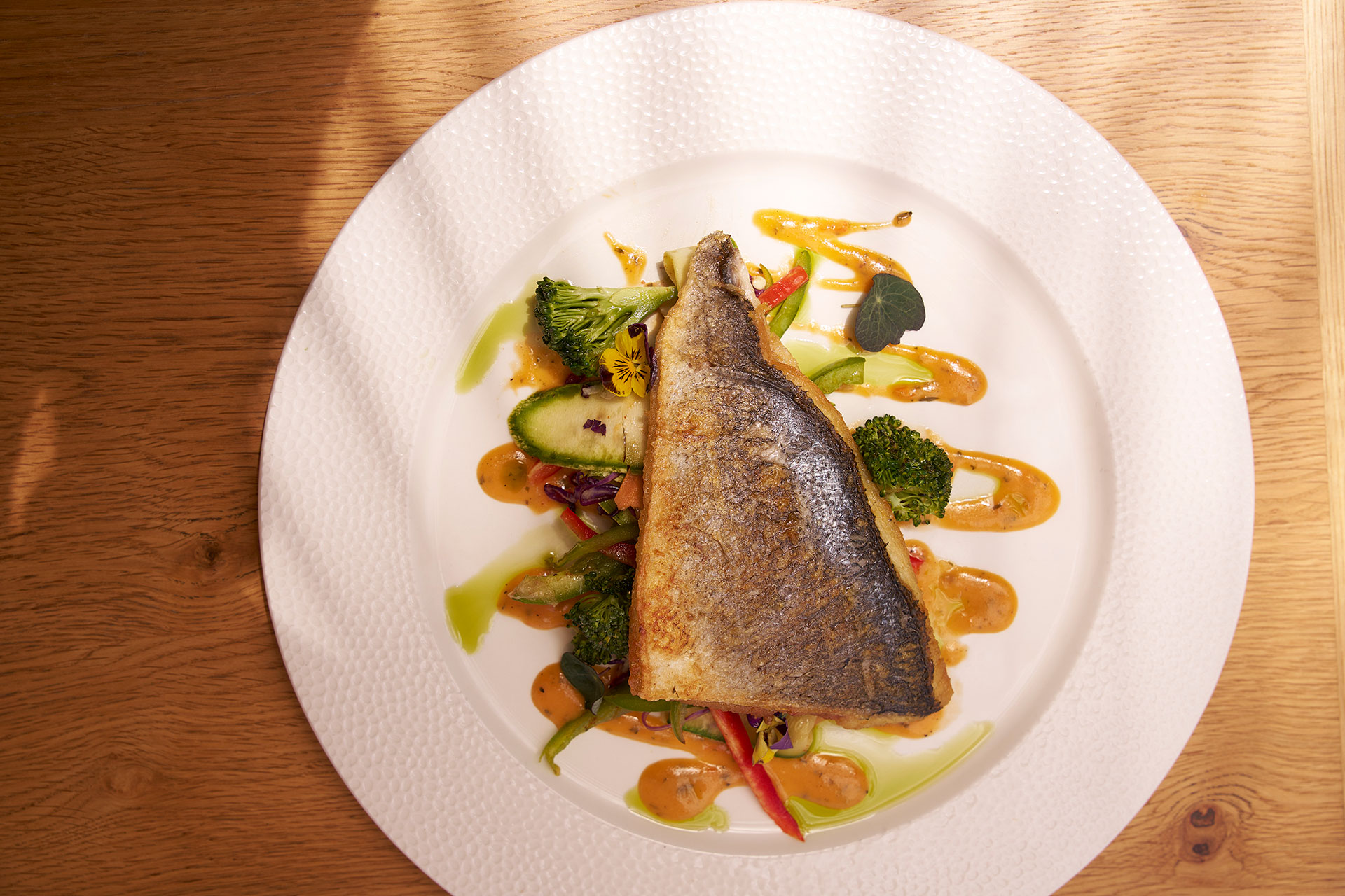 Seared fish fillet served over sautéed vegetables and drizzled with vibrant sauces on a white plate.