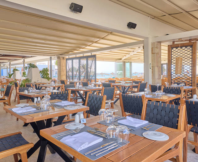 Open steakhouse dining area with wooden tables, black woven chairs, and views of the sea through glass panels