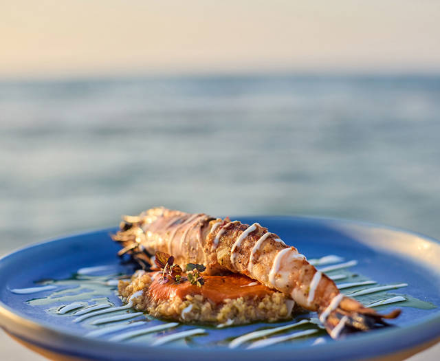 Grilled prawn elegantly plated on a blue dish with sauce and grains, set against a seaside backdrop.