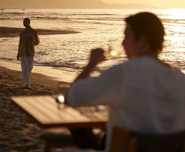Man drinking at a beachside table while woman walks along the shore at sunset