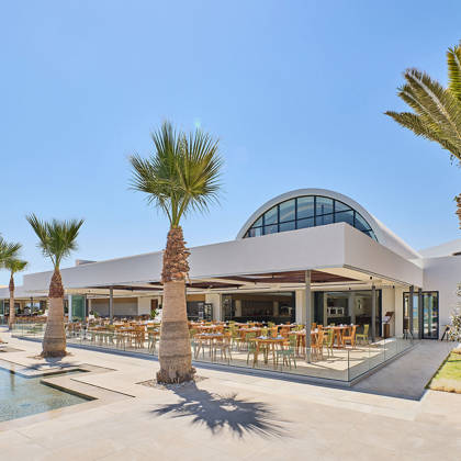 Exterior view of a modern open-air restaurant with palm trees in front and arched glass architecture above.