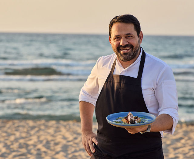 Smiling chef in uniform holding a plated seafood dish on the beach at sunset.