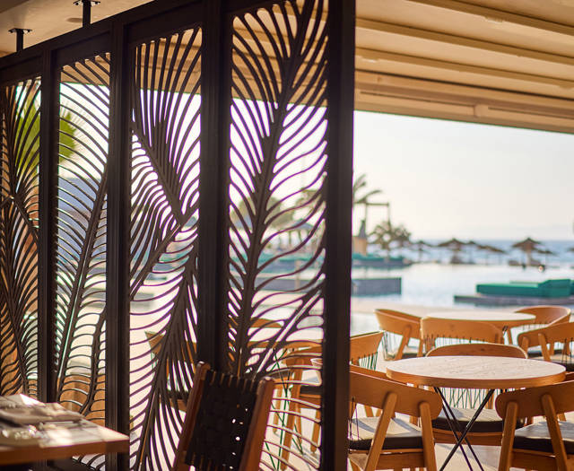 Decorative wooden screen with leaf patterns separating a steakhouse dining area from a seaside terrace with wooden tables and chairs.