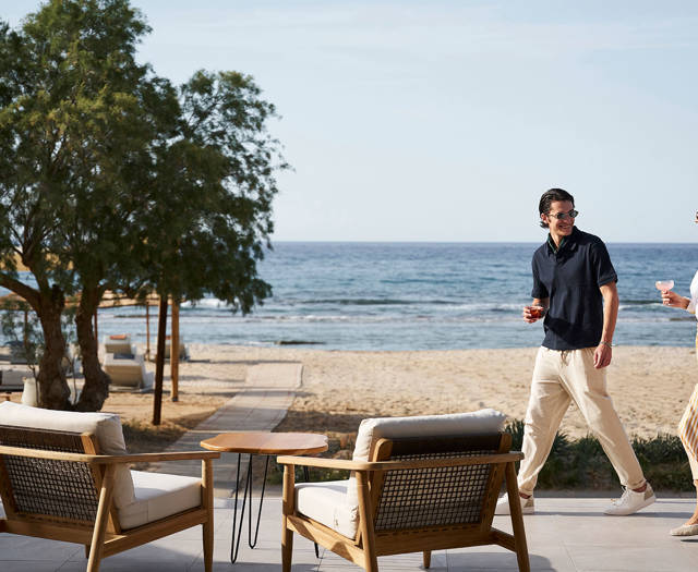 Couple walking along a seaside patio with drinks, passing between cushioned wooden chairs facing the ocean.