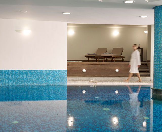 Guest in bathrobe walking by indoor spa pool with mosaic tiles.