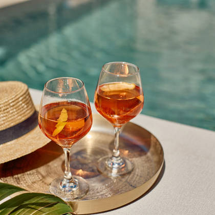 Two spritz cocktails on a tray beside a pool with a straw hat.